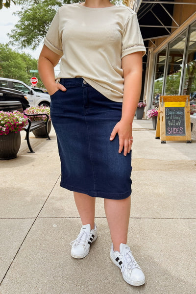 Woman wearing dark wash denim skirt with front and back pockets, subtle distressing, and classic button-and-zipper closure.