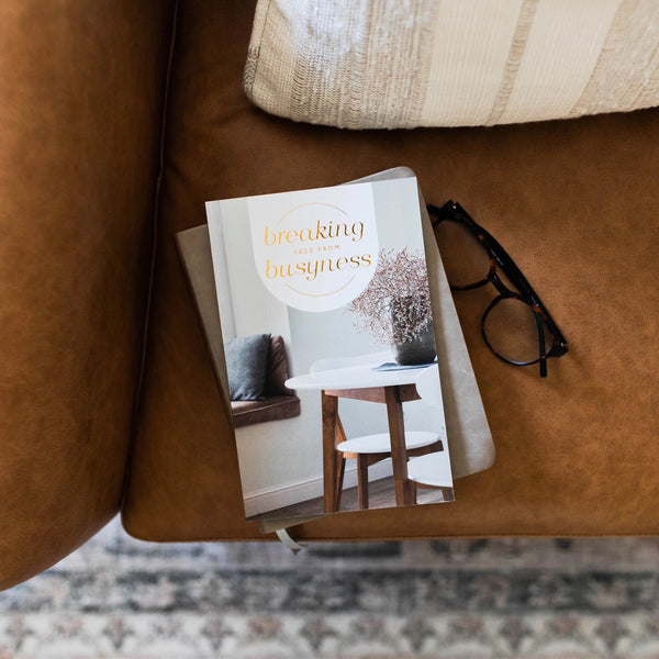 A booklet titled 'breaking busyness' is placed on a sofa next to a pair of glasses, indicating themes of quiet reflection and simplicity.