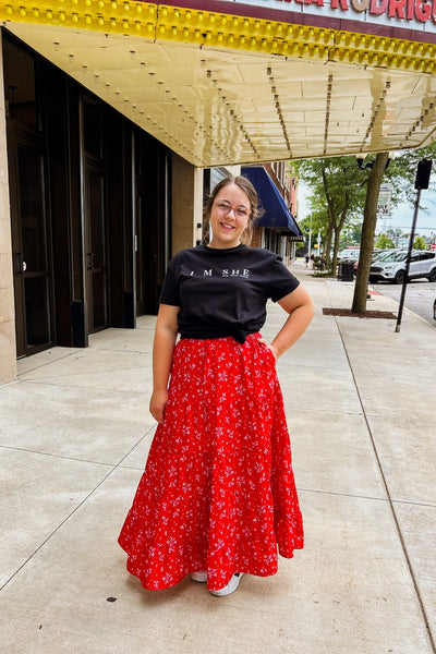 Woman wearing bright red floral maxi skirt with tiered design, elastic waist, drawstring, and side pockets. Styled with a black tee