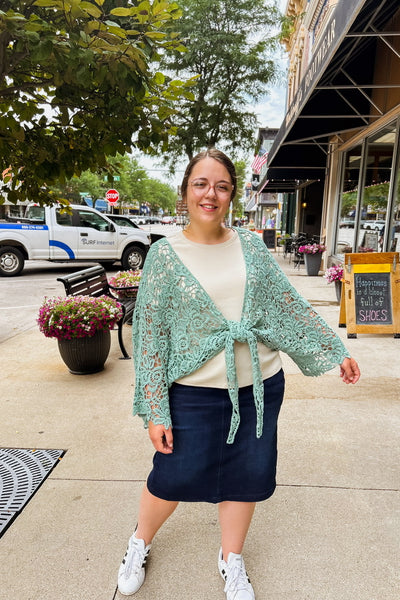 Woman wearing a mint crochet kimono with floral petal design, tied at the waist, layered over an oatmeal top and denim skirt