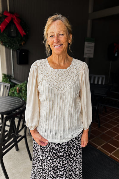 A woman standing outdoors wearing a cream-colored crochet sweater top with 3/4 sleeves and a key-hole neckline, paired with a floral skirt.