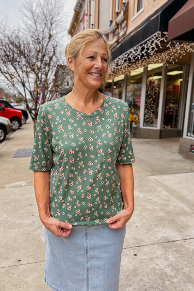 A woman standing on the sidewalk wearing a green floral print rib knit top with elbow length sleeves and a modest neckline, paired with a blue skirt.