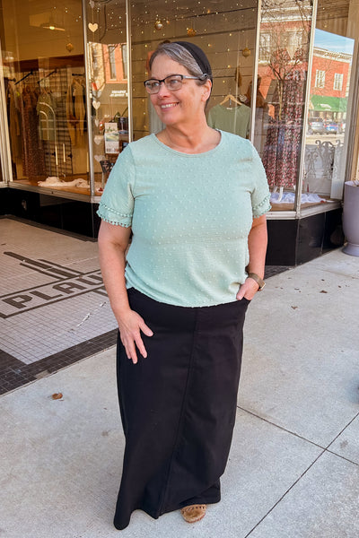 Woman wearing a long black denim skirt with comfort waistband, front and back pockets, styled with a green top
