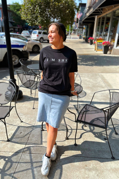 Woman wearing black graphic tee with “I M SHE, Strong. Honored. Esteemed.” on front and I M SHE logo on back, made of 100% cotton.