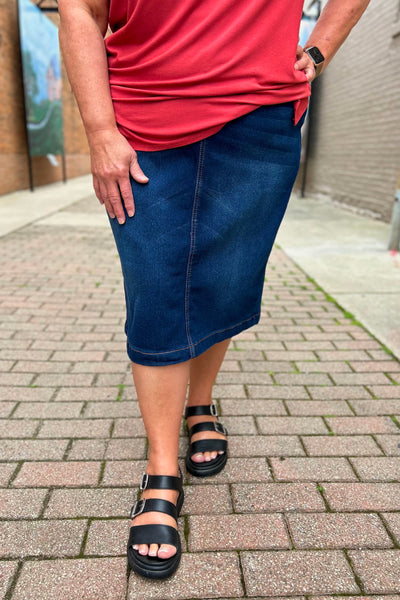 Woman wearing a dark wash midi denim skirt with comfort waistband, front and hip pockets, and 4-inch back slit.