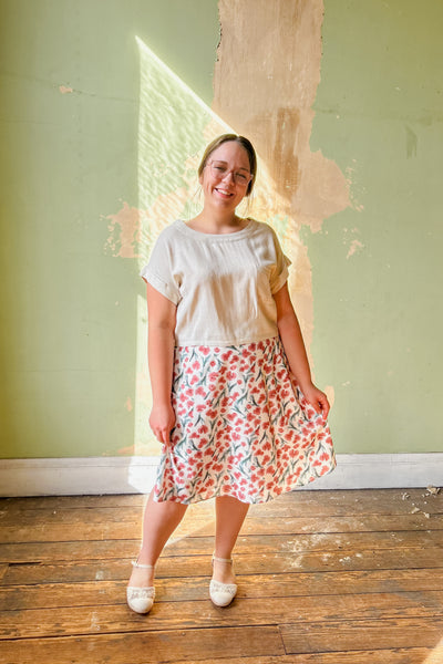 A woman standing in a room with natural lighting, wearing a midi floral skirt with a cream background and deep pink florals. She paired it with a linen top and white shoes.