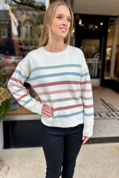 Young woman wearing a cozy white sweater with muted stripes, black jeans, and black booties, standing outside a boutique shop on a fall day.