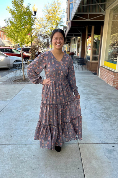 Woman standing on a downtown sidewalk wearing a long floral maxi dress with long sleeves, a V-neckline, and a tiered skirt. The dress has a smocked waist and cuffs, creating a soft, flowy silhouette. She is smiling and holding a portion of the dress hem, with fall trees and shopfronts in the background.