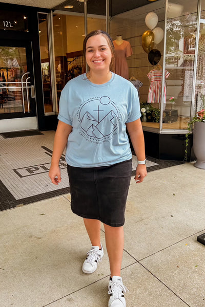 A person standing on the sidewalk wearing a light blue t-shirt with a “Faith Can Move Mountains” graphic, round-neck, and short sleeves, paired with a black skirt and white sneakers.