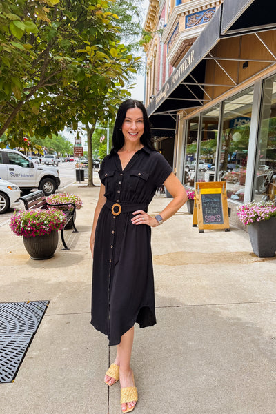 Woman wearing a black button-front midi dress with belt, curved hemline, and classic tailored silhouette.