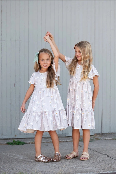 Two girls wearing matching taupe floral dresses with white flower prints and flutter sleeves.