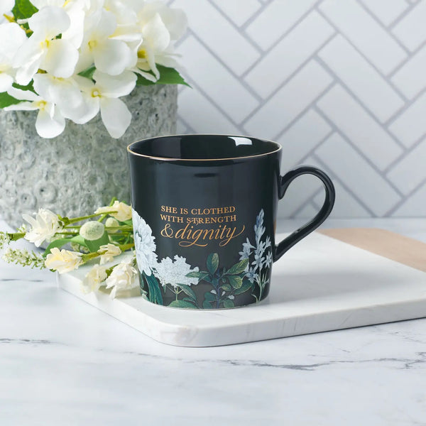 A black ceramic coffee mug with white floral patterns and text reading 'She is clothed with Strength & Dignity', placed on a white surface with a vase of flowers in the background.