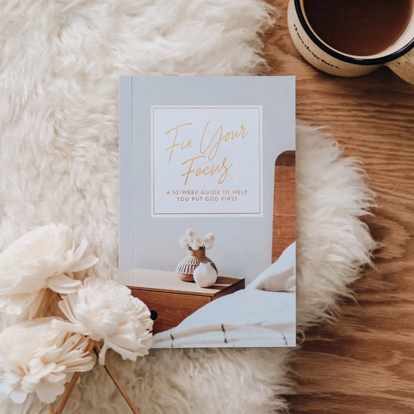 A book titled 'Fix Your Focus' with a white and gold cover, placed on a cozy white surface next to a cup of coffee and some decorative flowers.