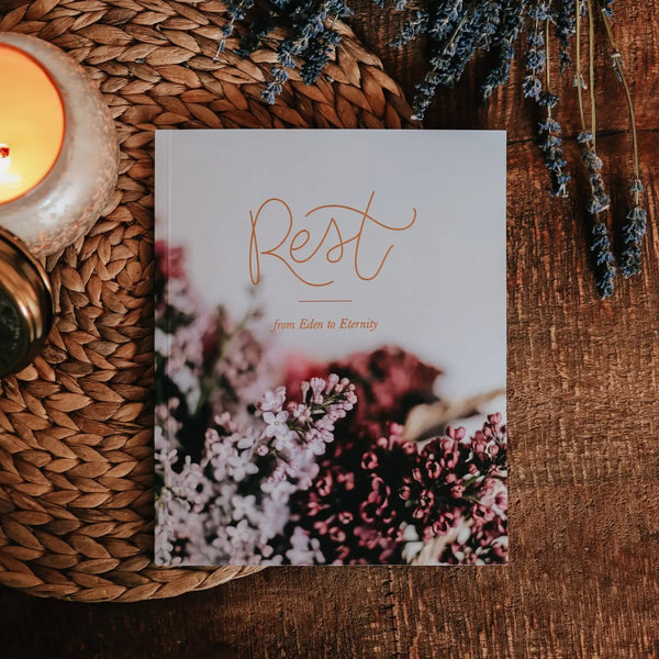 A book titled 'Rest' with a floral design on the cover, placed on a wicker table with a candle and lavender sprigs in the background.