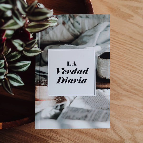 A book titled 'La Verdad Diaria | Daily Truth' is displayed with a background of a plant and a wooden surface. The book's cover is predominantly white with text, and it is presented as a vertical flip-through style.