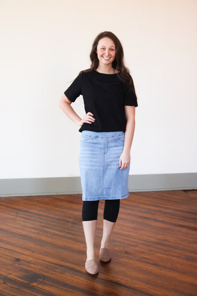 A person wearing a black top and black capri leggings under a midi denim skirt standing against a plain background