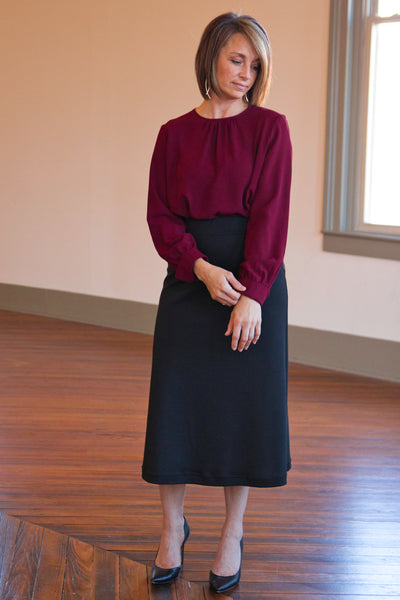 Woman wearing a knit black A-line midi skirt with a comfort waistband, paired with a red long-sleeve top, standing indoors.