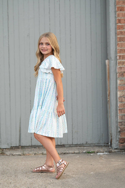 Young girl in a light blue dress standing against a gray wall.