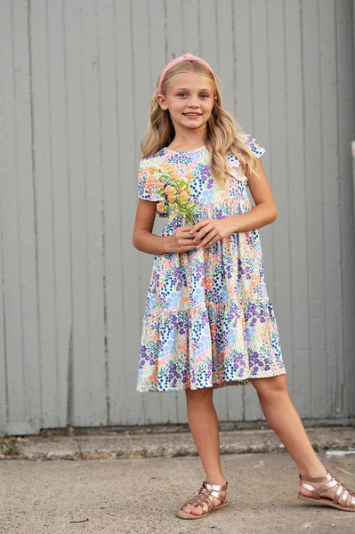 Young girl wearing a colorful floral dress standing against a gray wall.