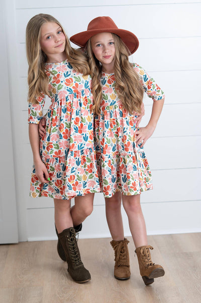 Two young girls wearing floral dresses and brown boots standing against a white wooden panel background.
