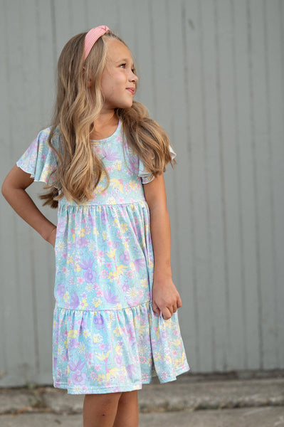 Little girl wearing a pastel floral dress with flutter sleeves and a tiered skirt in soft shades of lavender, blue, pink, and yellow, perfect for spring and summer.