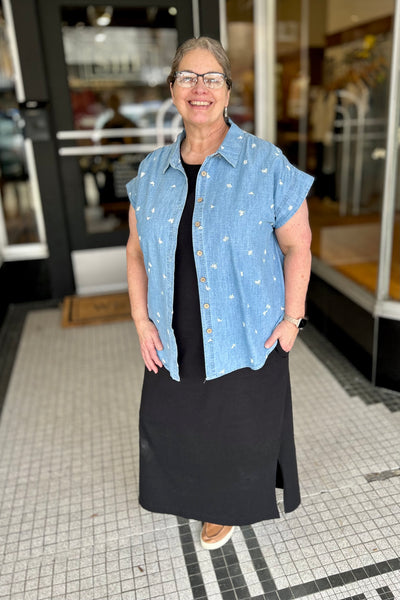 Woman wearing a embroidered chambray shirt over a black dress standing in front of a store entrance.