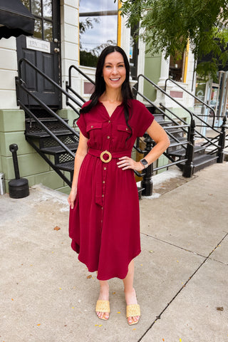 Woman wearing a burgundy button-front midi dress with belt, curved hemline, and classic tailored silhouette.
