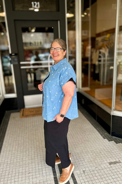 Person wearing a blue patterned shirt and black pants standing on a tiled floor in front of a building entrance.