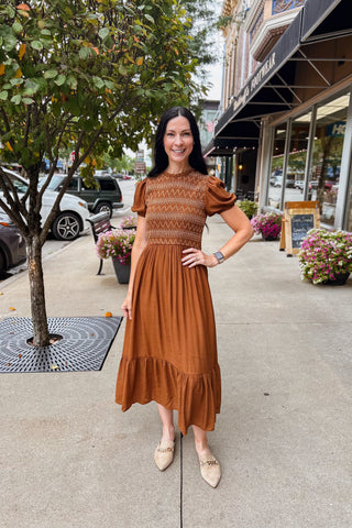 Woman wearing cinnamon brown smocked midi dress with puff sleeves, embroidered bodice, ruffle hem, and keyhole back closure.