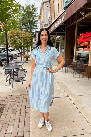 Woman wearing stripe chambray button-front dress with tie belt and shirt tail hem, styled with sneakers. 