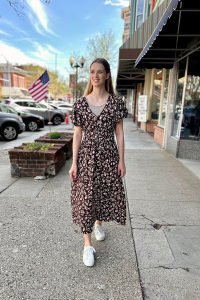 Woman in a floral dress standing on a sidewalk with storefronts and a flag in the background.