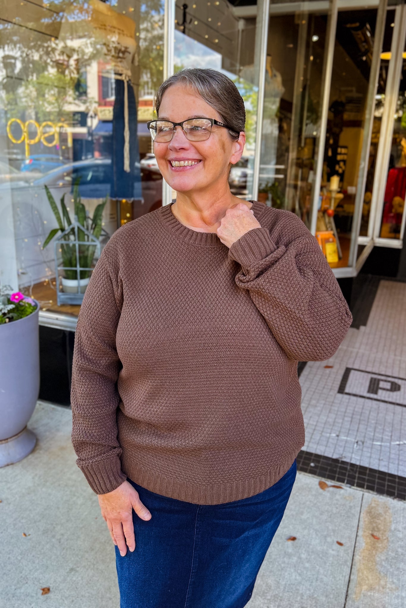 Woman on the side walk wearing a brown textured sweater with a flattering curved hemline. 