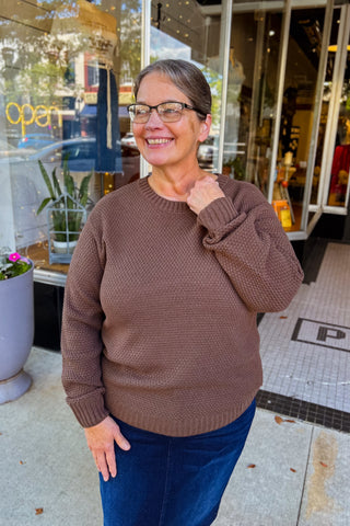 Woman on the side walk wearing a brown textured sweater with a flattering curved hemline. 