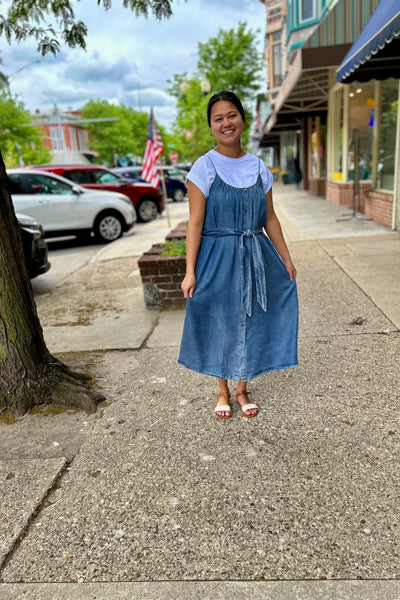 Woman in a blue denim dress standing on a sidewalk with storefronts and parked cars in the background.