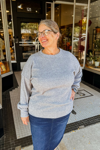 Lady  wearing a gray cozy light weight sweater with denim skirt  standing in front of I M SHE.