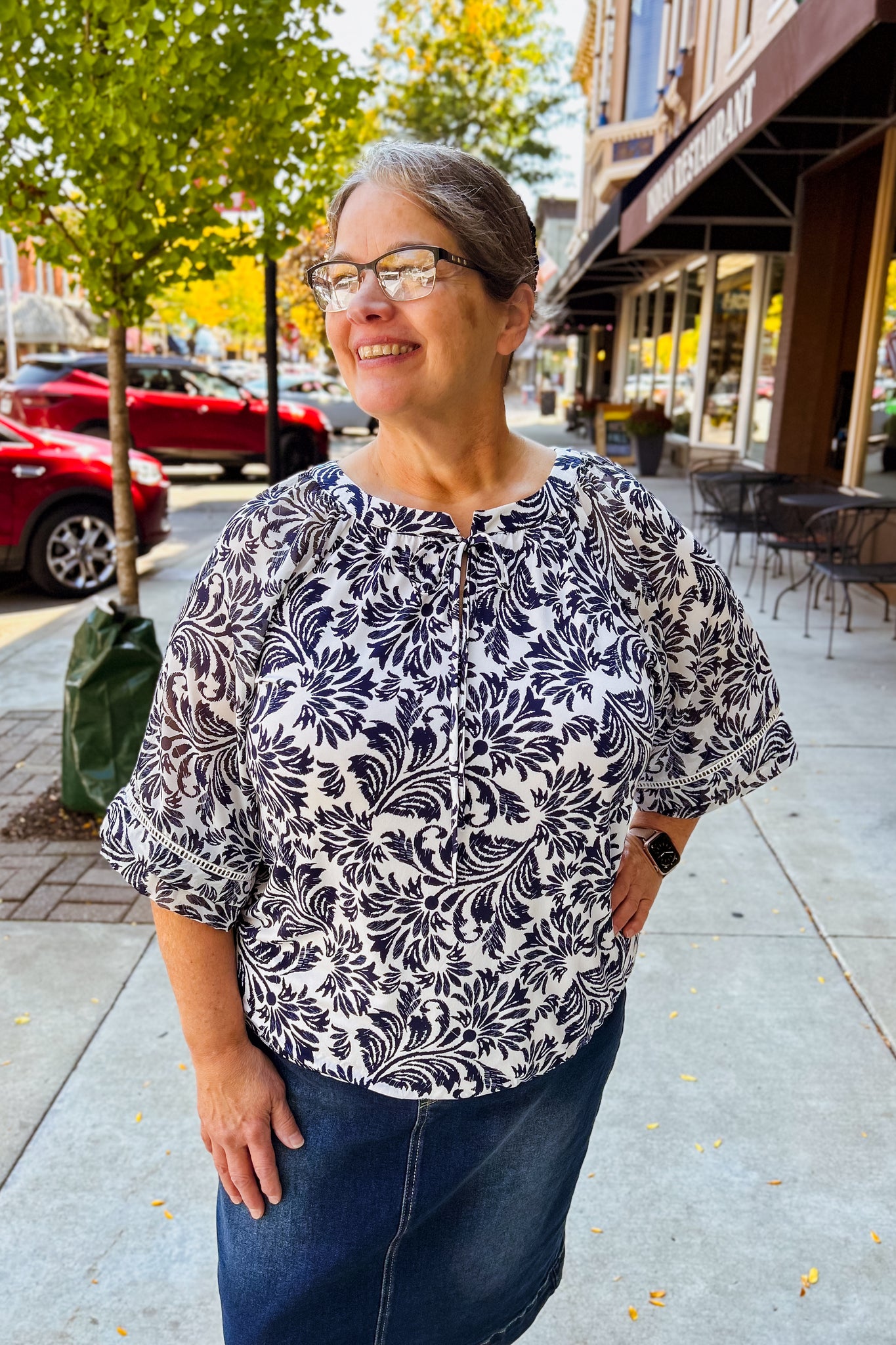 Woman wearing a navy  and white patterned blouse with 3/4 sleeves standing on a sidewalk.