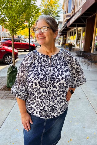 Woman wearing a navy  and white patterned blouse with 3/4 sleeves standing on a sidewalk.