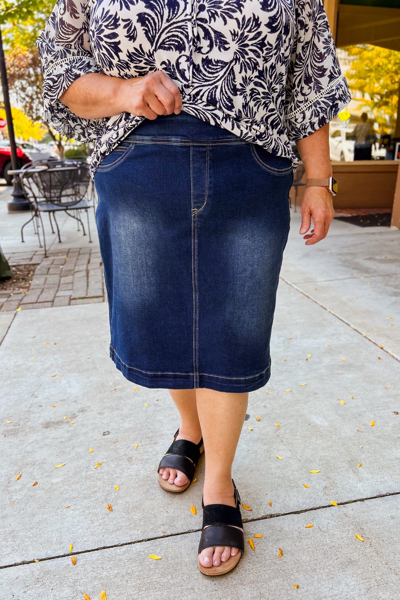 Person wearing a blue denim  knee length skirt and patterned top on a sidewalk.