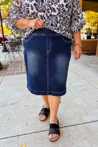 Person wearing a blue denim  knee length skirt and patterned top on a sidewalk.
