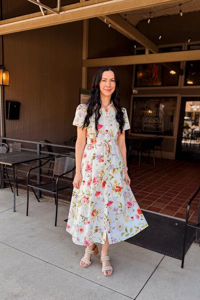 Woman in a modest floral shirt  dress standing outside a building with chairs and tables.