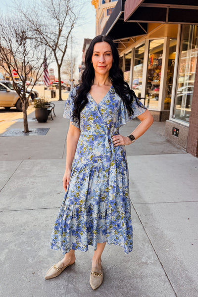 Woman in a blue floral dress standing on a sidewalk with storefronts in the background