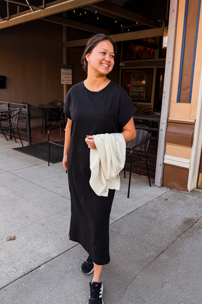 Woman in a black dress holding a white cloth, standing on a sidewalk with chairs and tables in the background.
