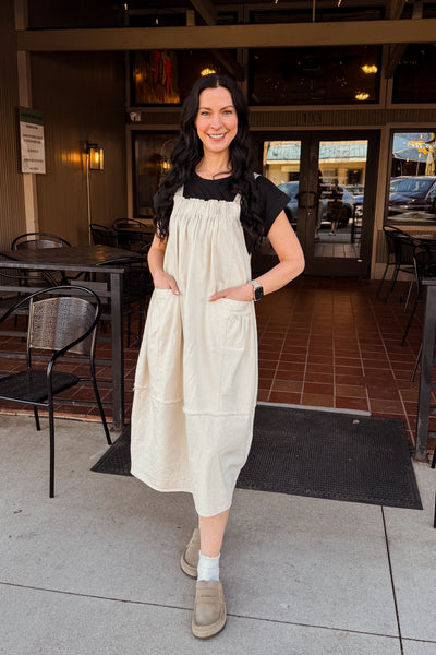 Woman in a modern modest oatmeal overall dress standing outside a restaurant with tables and chairs visible.