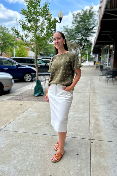 A woman stands on a city sidewalk wearing a short-sleeve olive green top with a subtle pattern, a white midi skirt, and brown slide sandals.