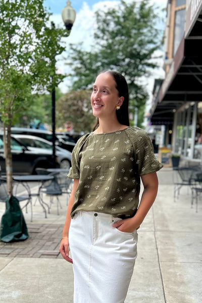 A woman standing on a city sidewalk wearing an olive green short-sleeve top with a small white floral pattern and white pants.