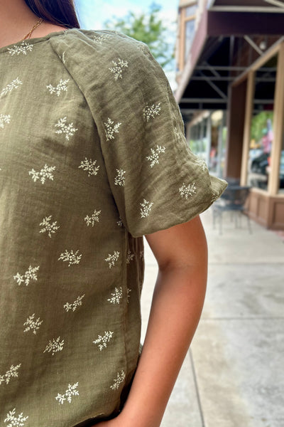 A close-up of a person wearing an olive green short-sleeve top with small white floral embroidery, photographed outdoors on a sidewalk.