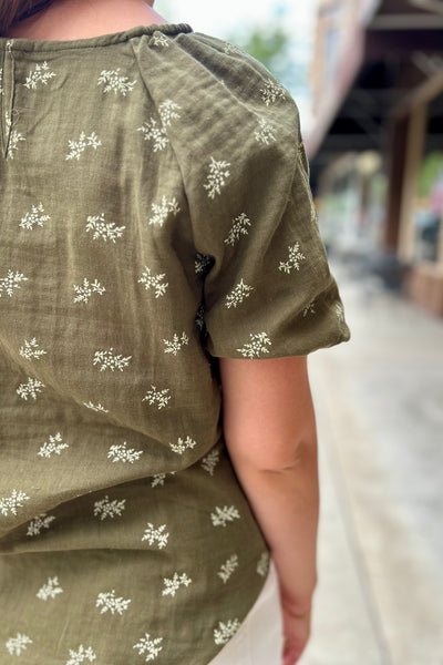 A close-up of a person wearing an olive green linen top with small white floral prints, photographed outdoors on a blurred walkway.