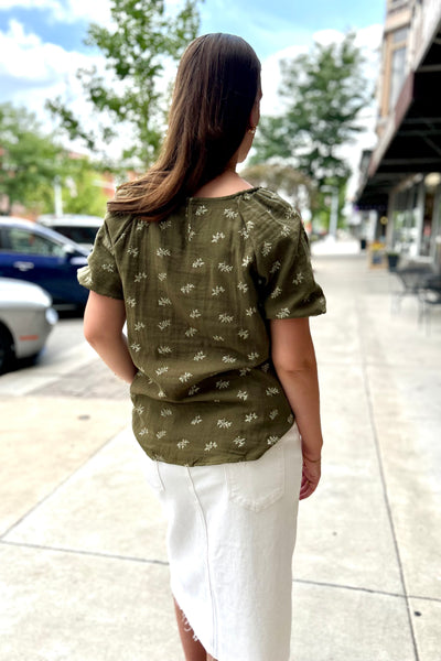 A woman viewed from behind wearing an olive green short-sleeve blouse with a small white floral pattern, paired with a white skirt, standing on a city sidewalk.