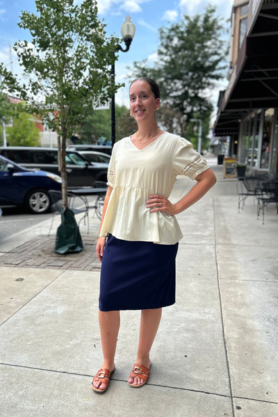 A woman modeling a cream-colored short-sleeve peplum top with lace-up sleeves and a navy blue knee-length skirt, standing on a city sidewalk.