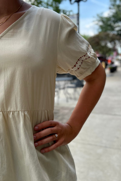 A close-up of a person wearing a cream-colored short-sleeve dress outdoors. The top has a V-neckline, a gathered waist seam, and delicate lace trim detailing on the sleeve cuffs.